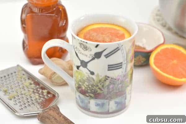 Citrus Spice Detox Tea Recipe for Gut Cleansing and Energy 3 Photo shows a mug or orange detox tea sitting on a counter top. An orange slice is floating in the mug. Nearby is a grater with ginger on it, a bottle of honey, and half a sliced orange.