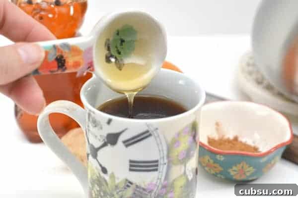 Citrus Spice Detox Tea Recipe for Gut Cleansing and Energy 5 Photo shows a mug of orange detox tea at the foreground of the image, with a small ceramic spoon of honey being poured into it by a hand.
