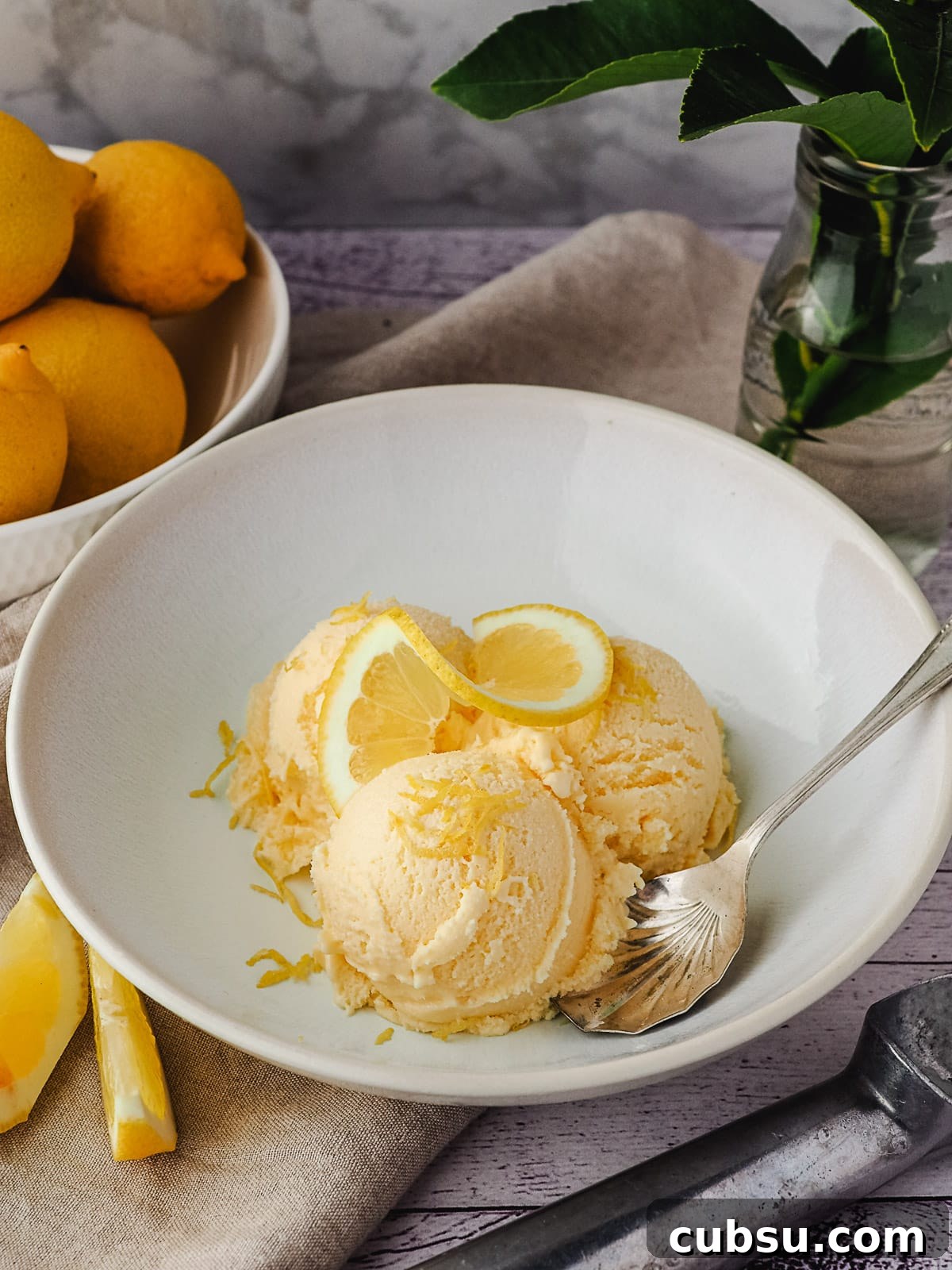 Creamy Lemon Ice Cream Recipe: Bright, Tangy Homemade Treat 12 Three scoops of ice cream an a bowl, garnished with fresh lemon, with lemons and ice cream scoop in background.