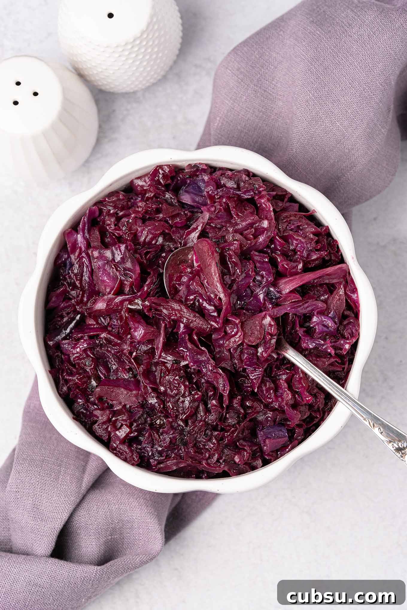 Slow-Braised Red Cabbage with Apple and Balsamic Glaze 2 An overhead view of a serving bowl of braised red cabbage with a spoon in it, on top of a linen napkin with white ceramic salt and pepper shakers behind it and to the left.
