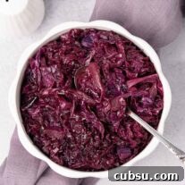 An overhead view of a serving bowl of braised red cabbage with a spoon in it, on top of a linen napkin with white ceramic salt and pepper shakers behind it and to the left.