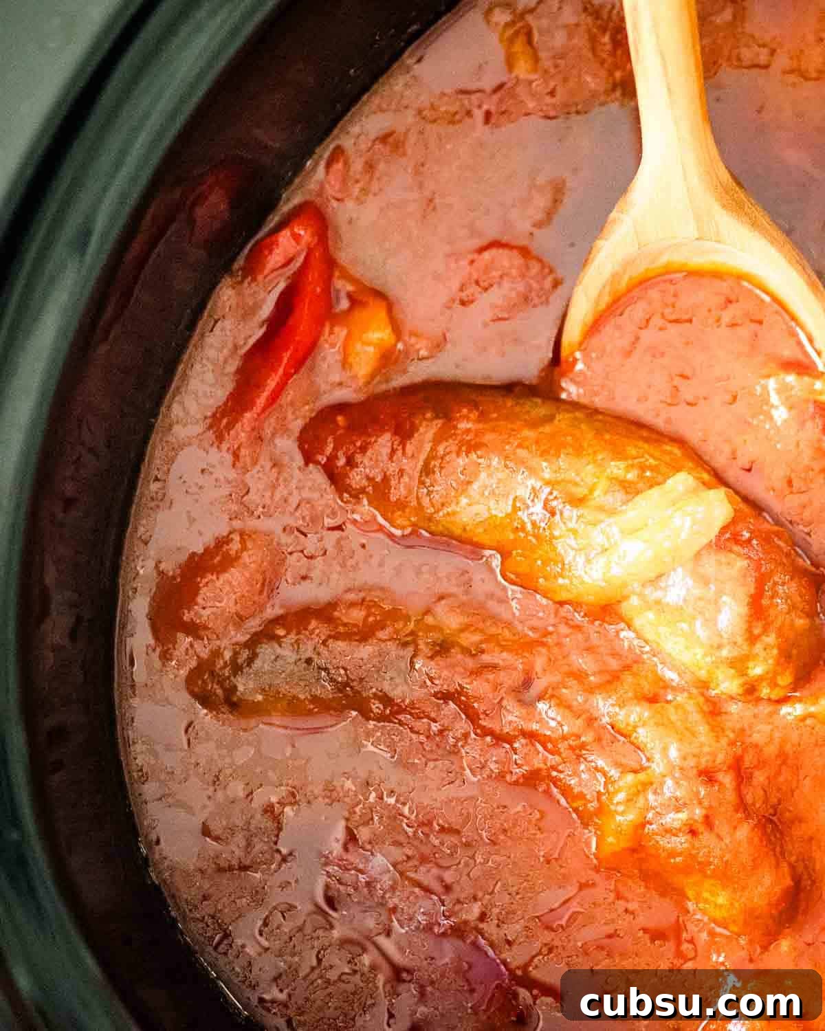 A wooden spoon in a slow cooker with sausage, peppers, and marinara sauce.