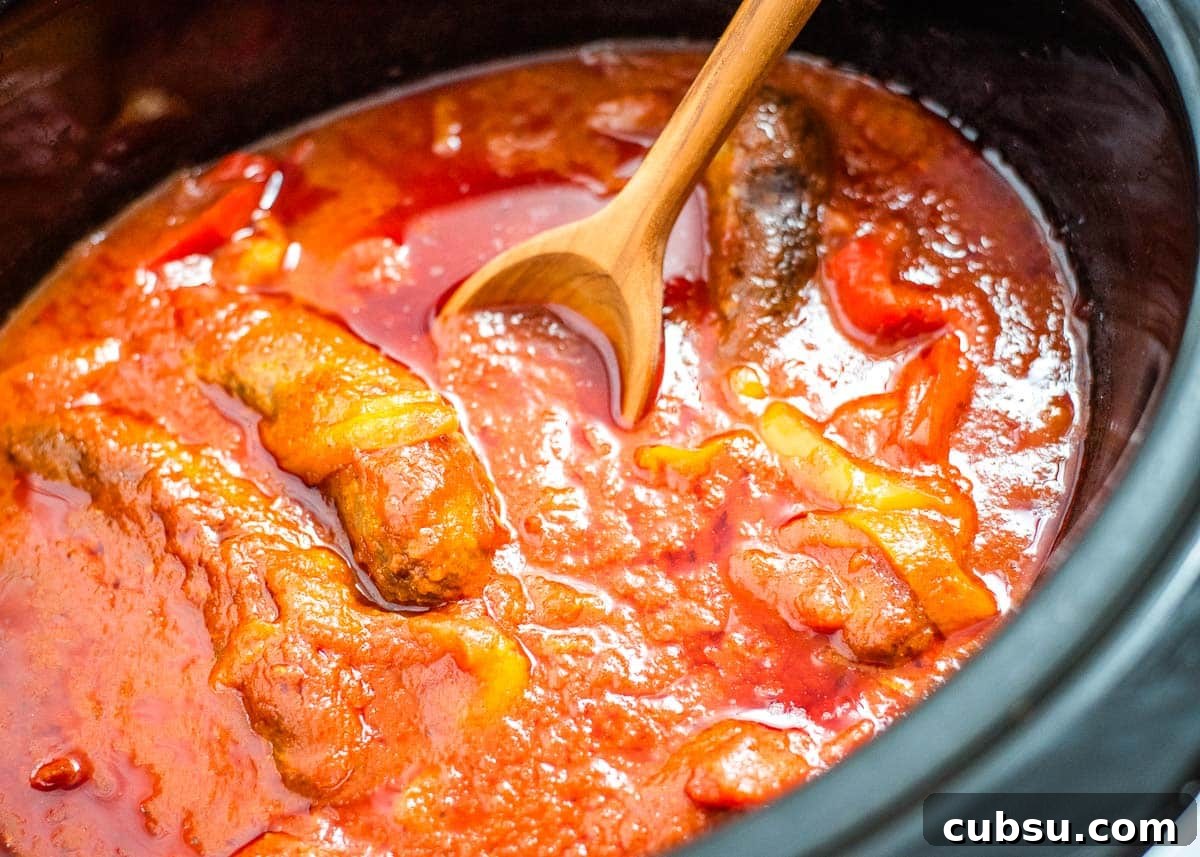 A wooden spoon in a slow cooker with sausage, peppers, and marinara sauce.