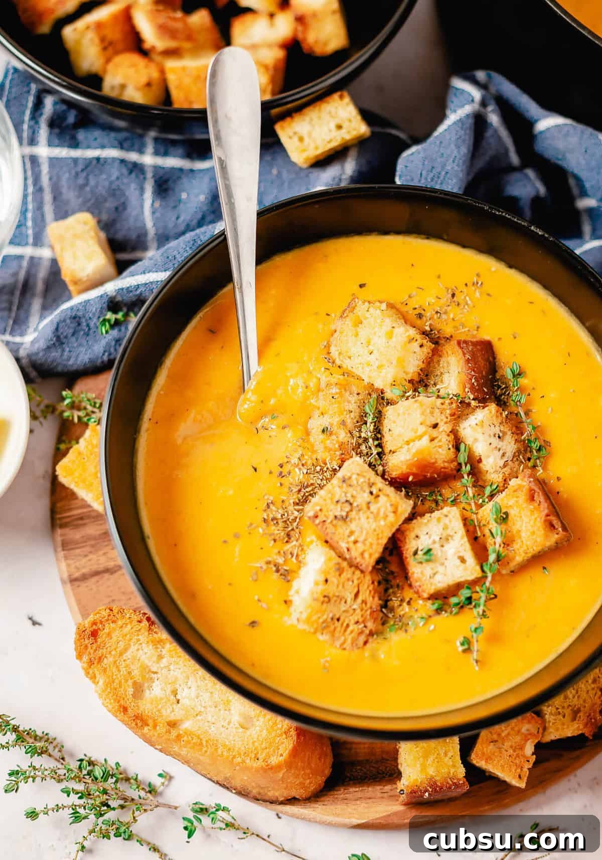 Butternut squash and sweet potato soup in a black bowl, topped with croutons, fresh thyme, and a spoon, with bread slices and a blue cloth in the background.
