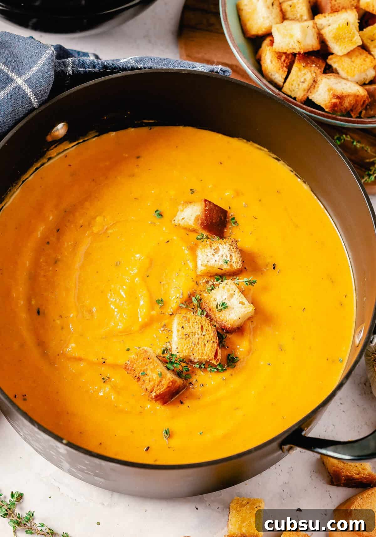 A pot of smooth butternut squash and sweet potato soup with croutons and fresh thyme on top, next to a small bowl of additional croutons.