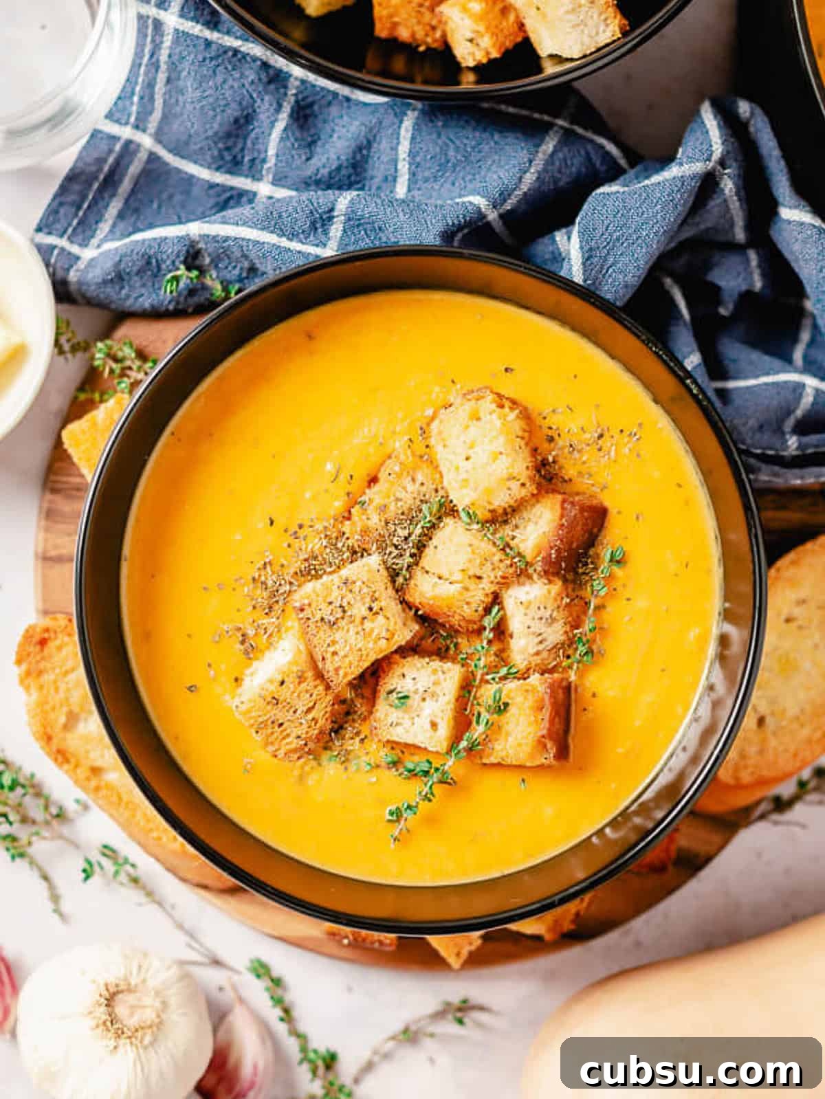 Butternut squash and sweet potato soup in a black bowl, topped with crispy croutons and a sprig of thyme, next to a bowl of additional croutons.