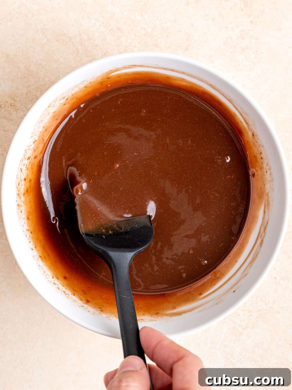 Stirring a melted chocolate cream mixture in a bowl with a small rubber spatula.