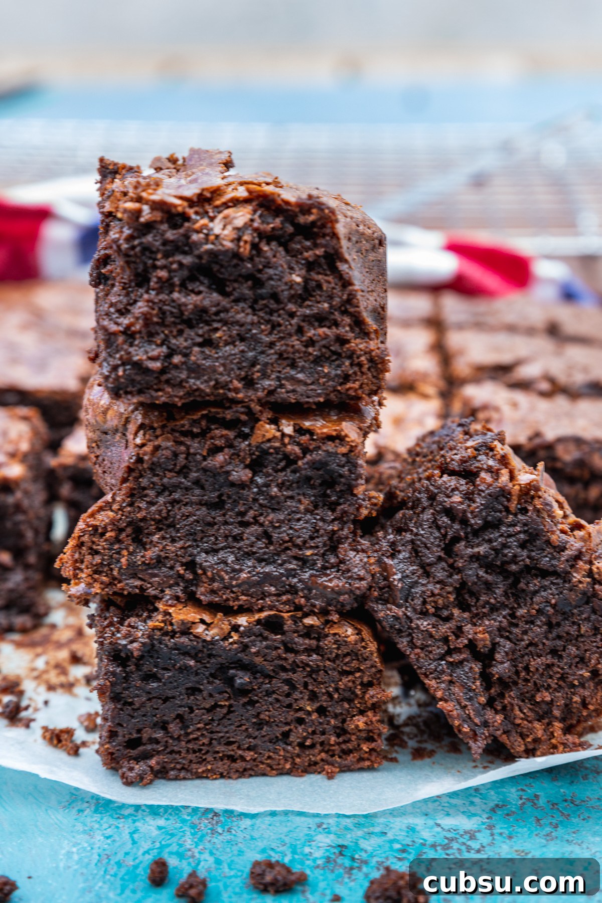One-Bowl Gluten-Free Brownies: Fudgy Recipe for Chocolate Lovers 8 A stack of 3 gluten-free brownies on a blue background and with the rest of the pan in the background