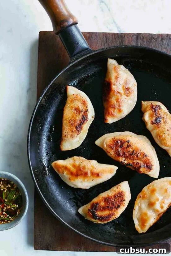 A black skillet with seven pork dumplings in it on a wooden surface and a cup of dipping sauce beside it.
