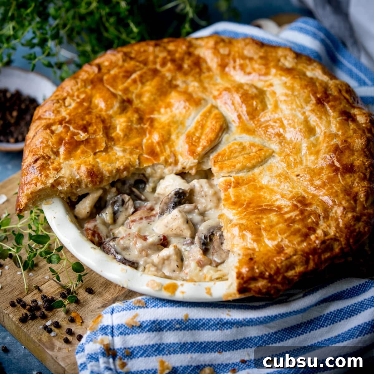 Golden-brown chicken and mushroom pie with a flaky pastry, one slice missing, showing creamy filling; on striped cloth with herbs and peppercorns.