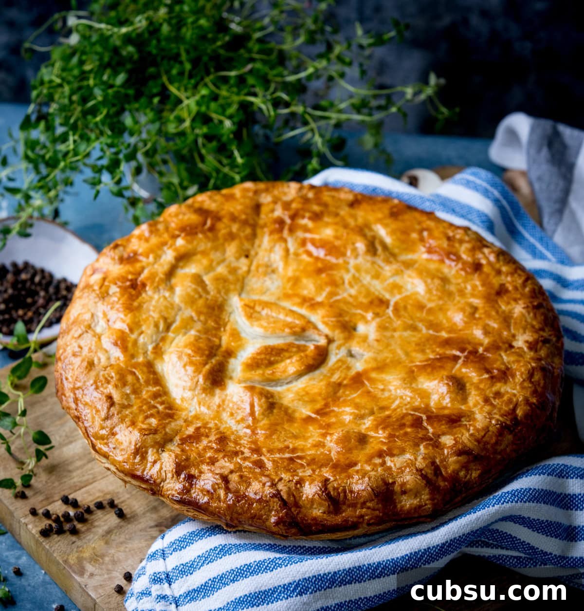 A golden-brown chicken and mushroom pie with a leaf design on top sits on a blue-striped kitchen tea towel, with herbs and peppercorns nearby.