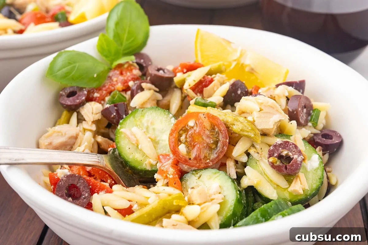 Mediterranean Tuna Orzo Salad with Olives, Feta & Lemon 2 A bowl of Mediterranean tuna orzo salad with sliced cucumbers, cherry tomatoes, olives, herbs, and a lemon wedge. A fork is placed inside the bowl.