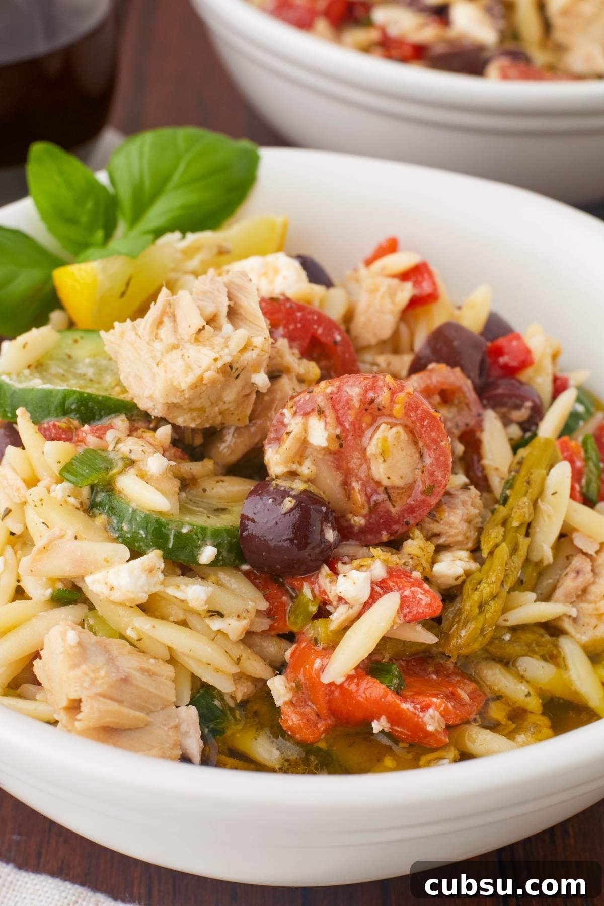 Mediterranean Tuna Orzo Salad with Olives, Feta & Lemon 11 A white bowl filled with Mediterranean tuna orzo salad, featuring cherry tomatoes, olives, feta cheese, and fresh basil leaves.