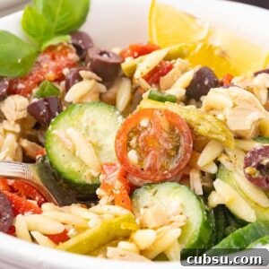 A white bowl brimming with a Mediterranean tuna orzo salad features cherry tomatoes, cucumber slices, olives, and basil, all crowned with a lemon wedge. A fork rests beside the vibrant mix.