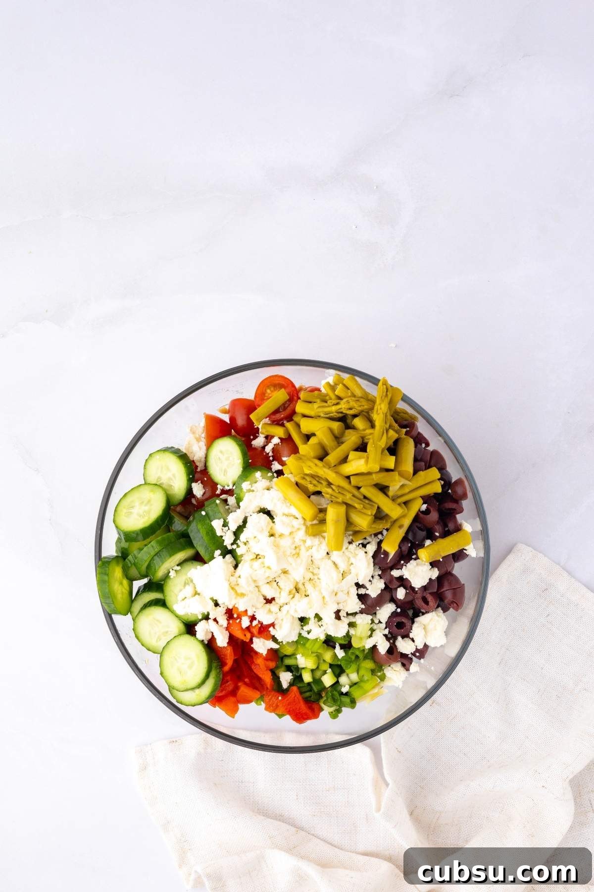 Mediterranean Tuna Orzo Salad with Olives, Feta & Lemon 15 A bowl of Mediterranean tuna orzo salad mixed with cucumbers, red peppers, olives, green beans, scallions, feta cheese, and cherry tomatoes sits on a white surface beside a beige napkin.