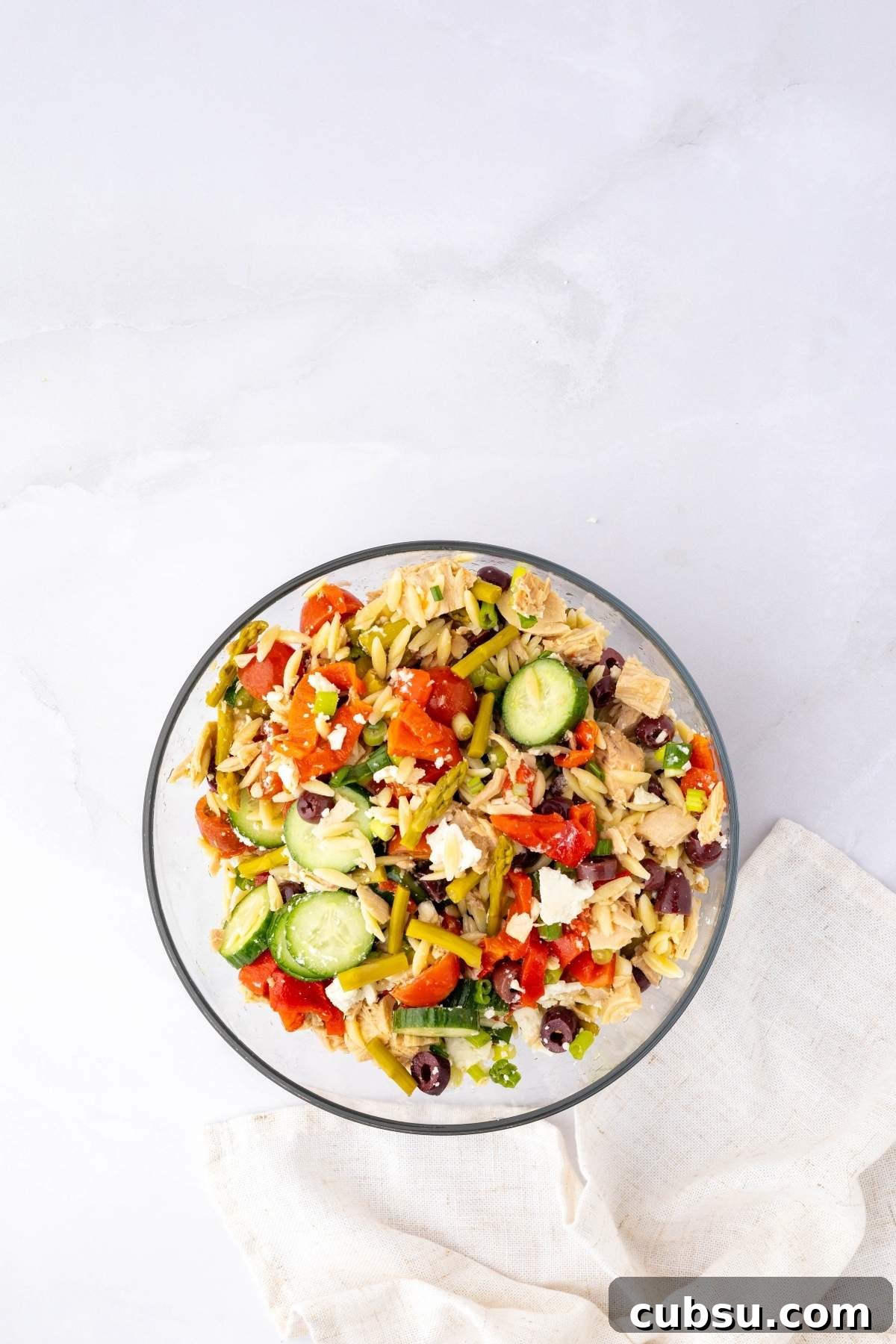 Mediterranean Tuna Orzo Salad with Olives, Feta & Lemon 16 A glass bowl brimming with Mediterranean tuna orzo salad, featuring cucumbers, cherry tomatoes, olives, feta cheese, and greens, is elegantly placed on a white cloth.