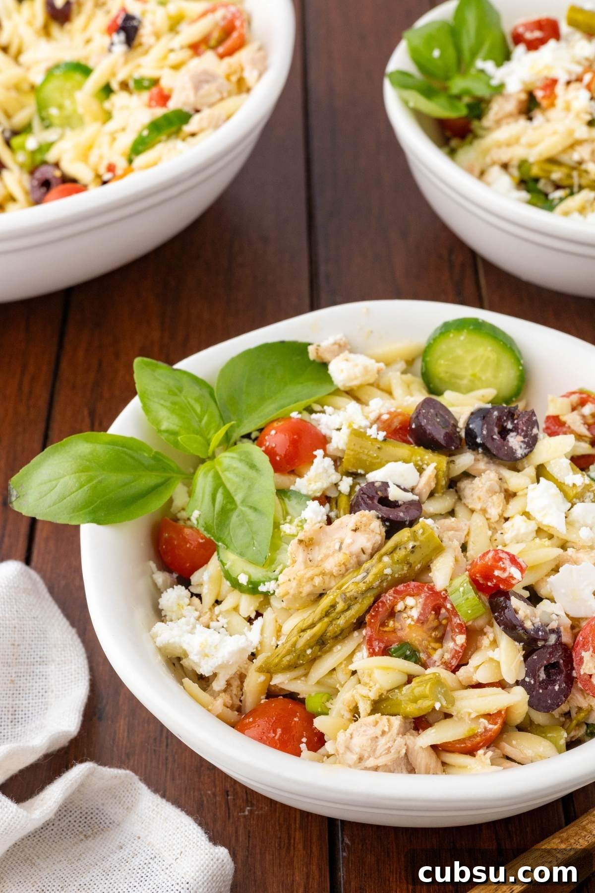 Mediterranean Tuna Orzo Salad with Olives, Feta & Lemon 19 A bowl of Mediterranean tuna orzo salad with cherry tomatoes, cucumber, olives, asparagus, feta cheese, and basil leaves sits on a wooden table. Two similar bowls are in the background.