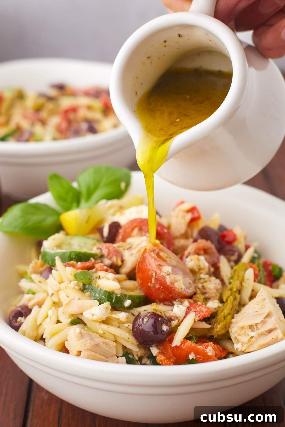 Mediterranean Tuna Orzo Salad with Olives, Feta & Lemon 4 A hand pours dressing onto a Mediterranean tuna orzo salad, complete with vibrant vegetables, olives, and freshly garnished basil.