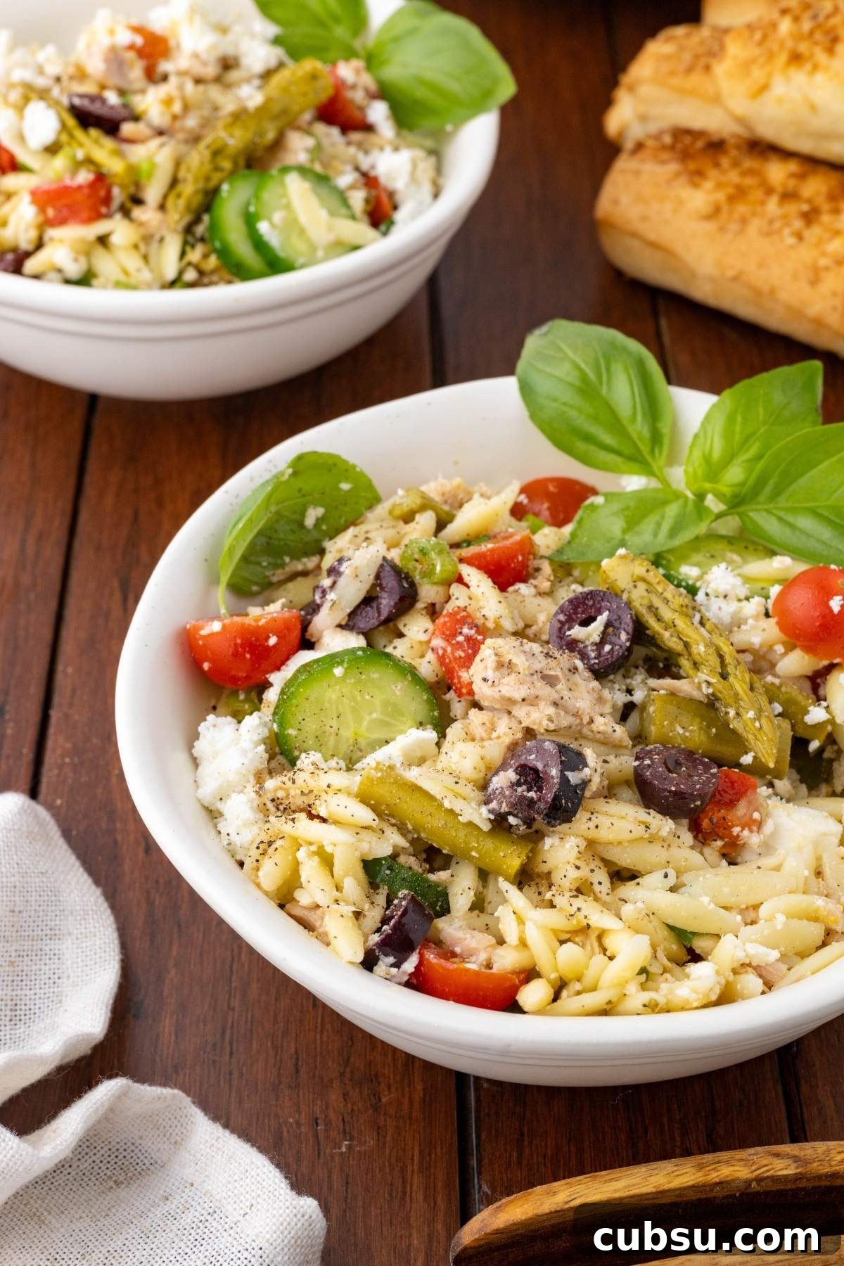 Mediterranean Tuna Orzo Salad with Olives, Feta & Lemon 5 Two bowls of Mediterranean tuna orzo salad with cherry tomatoes, cucumbers, olives, feta cheese, and basil garnish on a wooden table, paired with breadsticks.