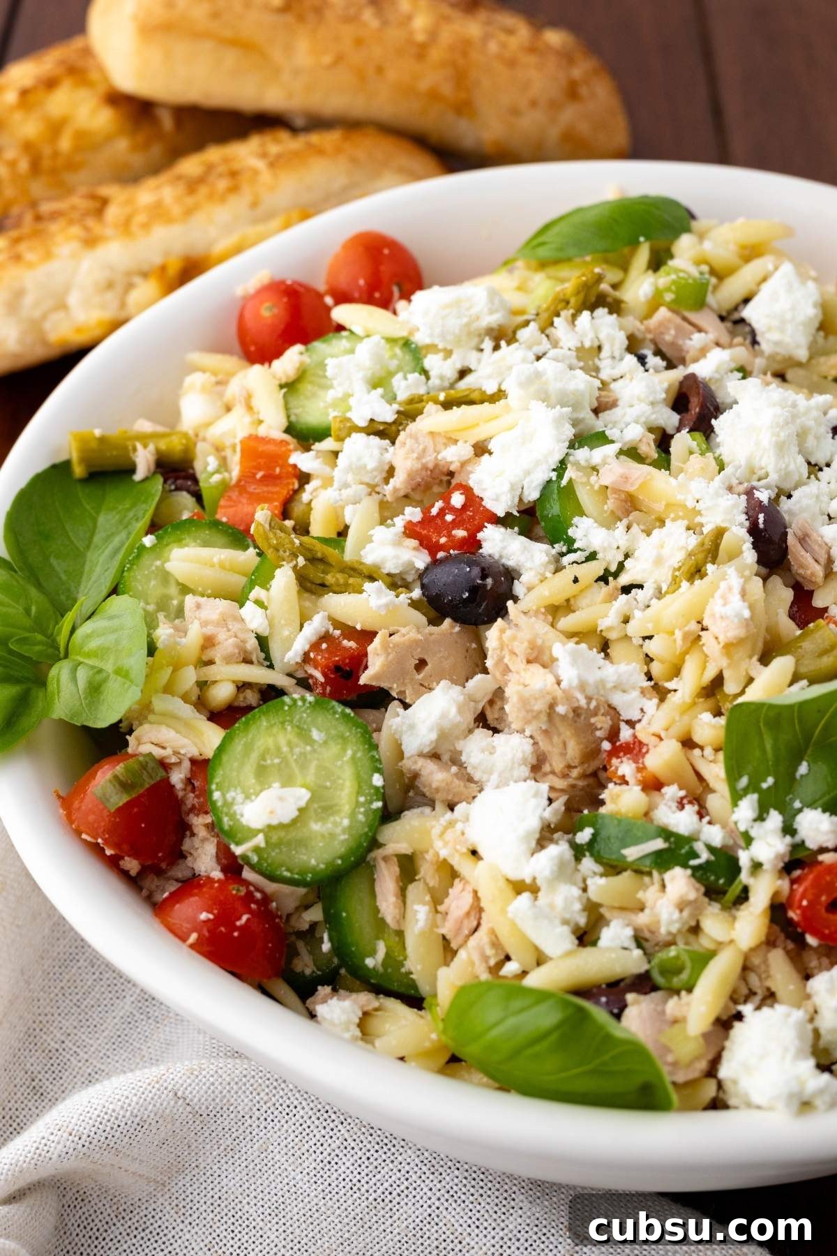 Mediterranean Tuna Orzo Salad with Olives, Feta & Lemon 7 A bowl of Mediterranean tuna orzo salad with feta, cherry tomatoes, cucumber, olives, and greens sits on a table next to two pieces of bread.