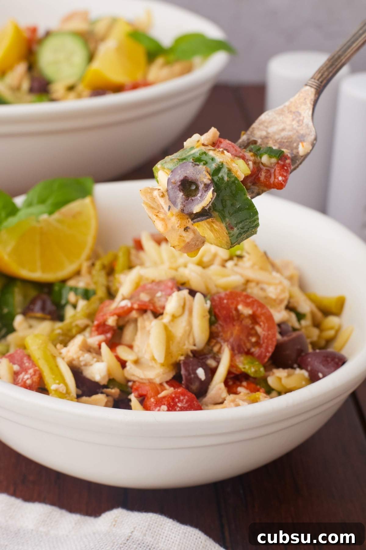 Mediterranean Tuna Orzo Salad with Olives, Feta & Lemon 8 A fork holds a bite of Mediterranean tuna orzo salad with chopped vegetables and olives over a bowl of the same salad, garnished with lemon slices and basil.