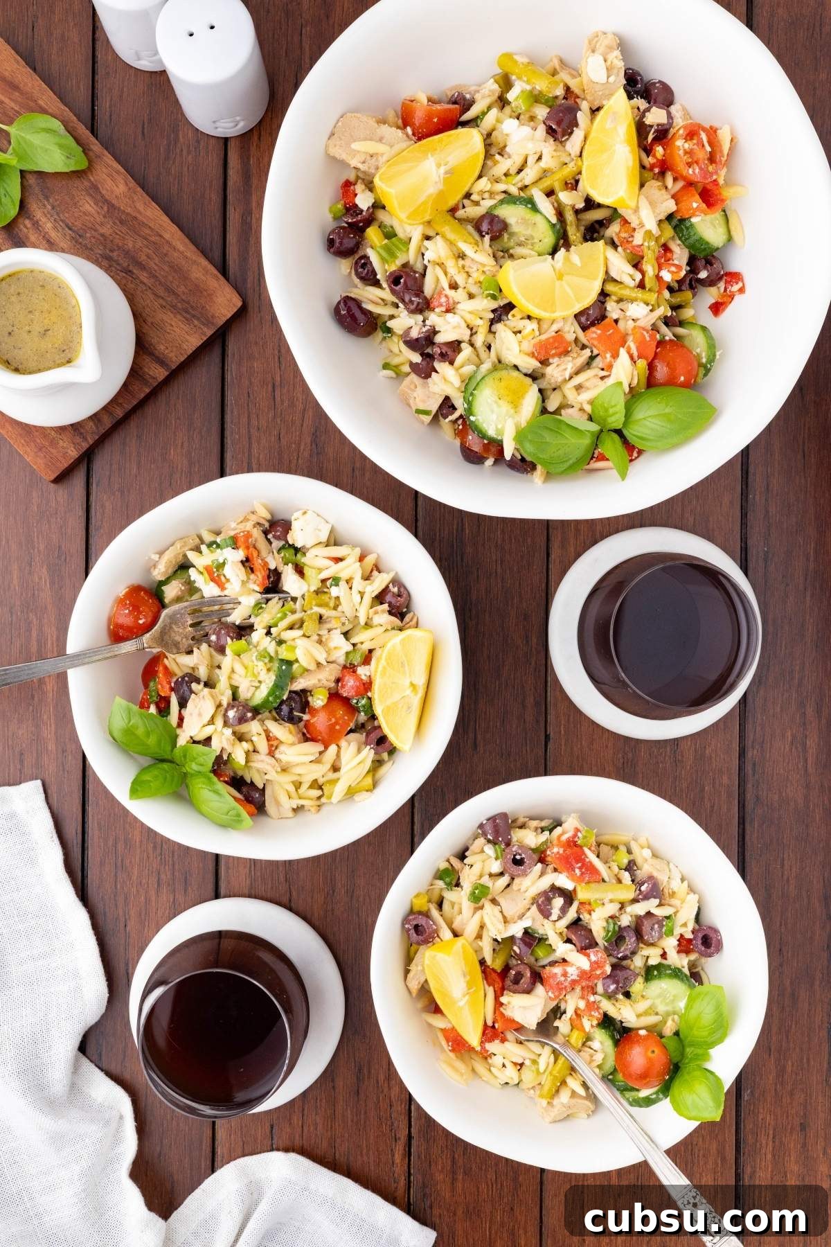 Mediterranean Tuna Orzo Salad with Olives, Feta & Lemon 9 Three bowls of Mediterranean tuna orzo salad with lemon wedges, cherry tomatoes, olives, and greens on a wooden table, accompanied by two glasses of red wine and a small dish of dressing.