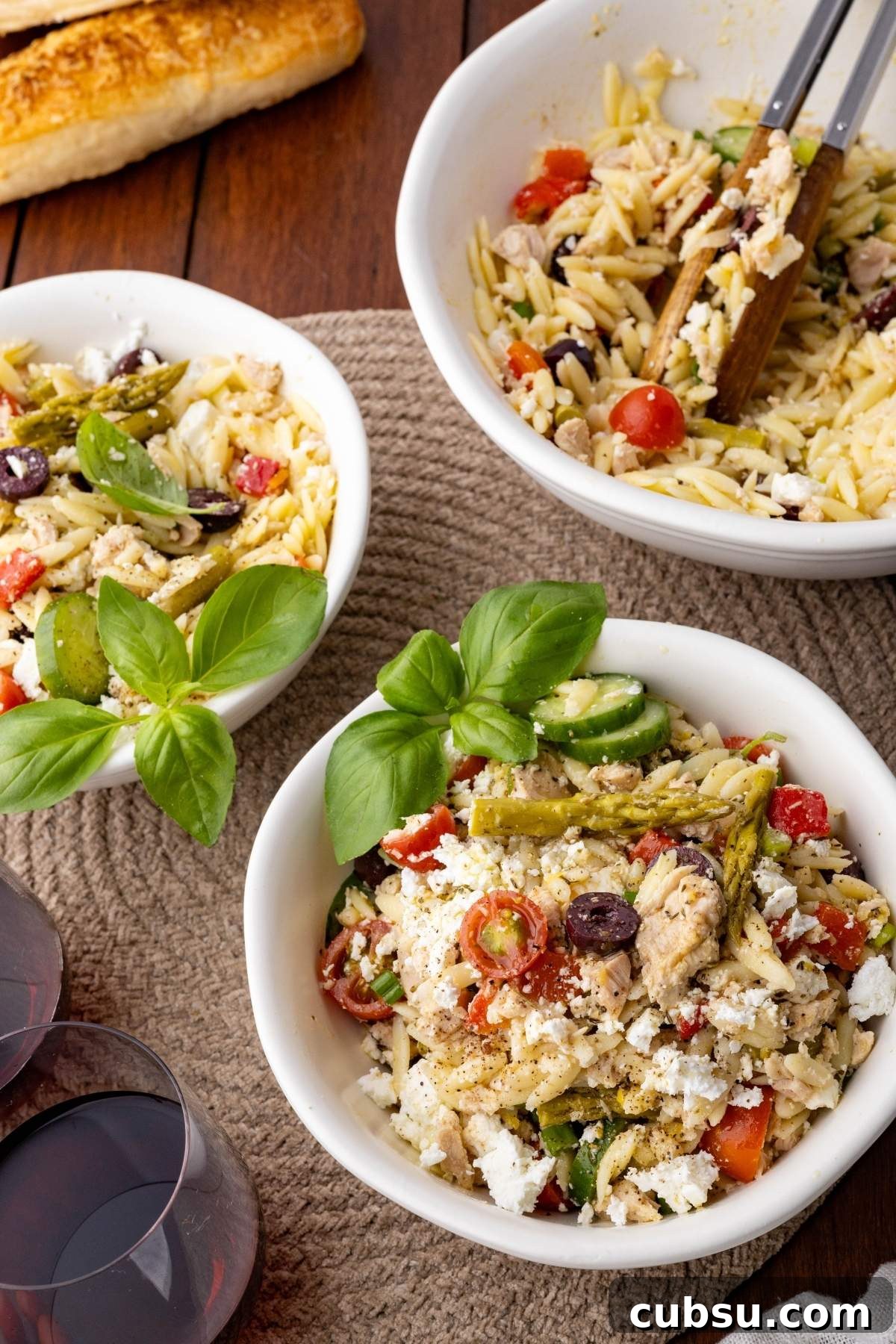 Mediterranean Tuna Orzo Salad with Olives, Feta & Lemon 10 Three bowls of Mediterranean tuna orzo salad with olives, tomatoes, zucchini, asparagus, and feta cheese are on a table with basil garnish, alongside two glasses of red wine and a loaf of bread.