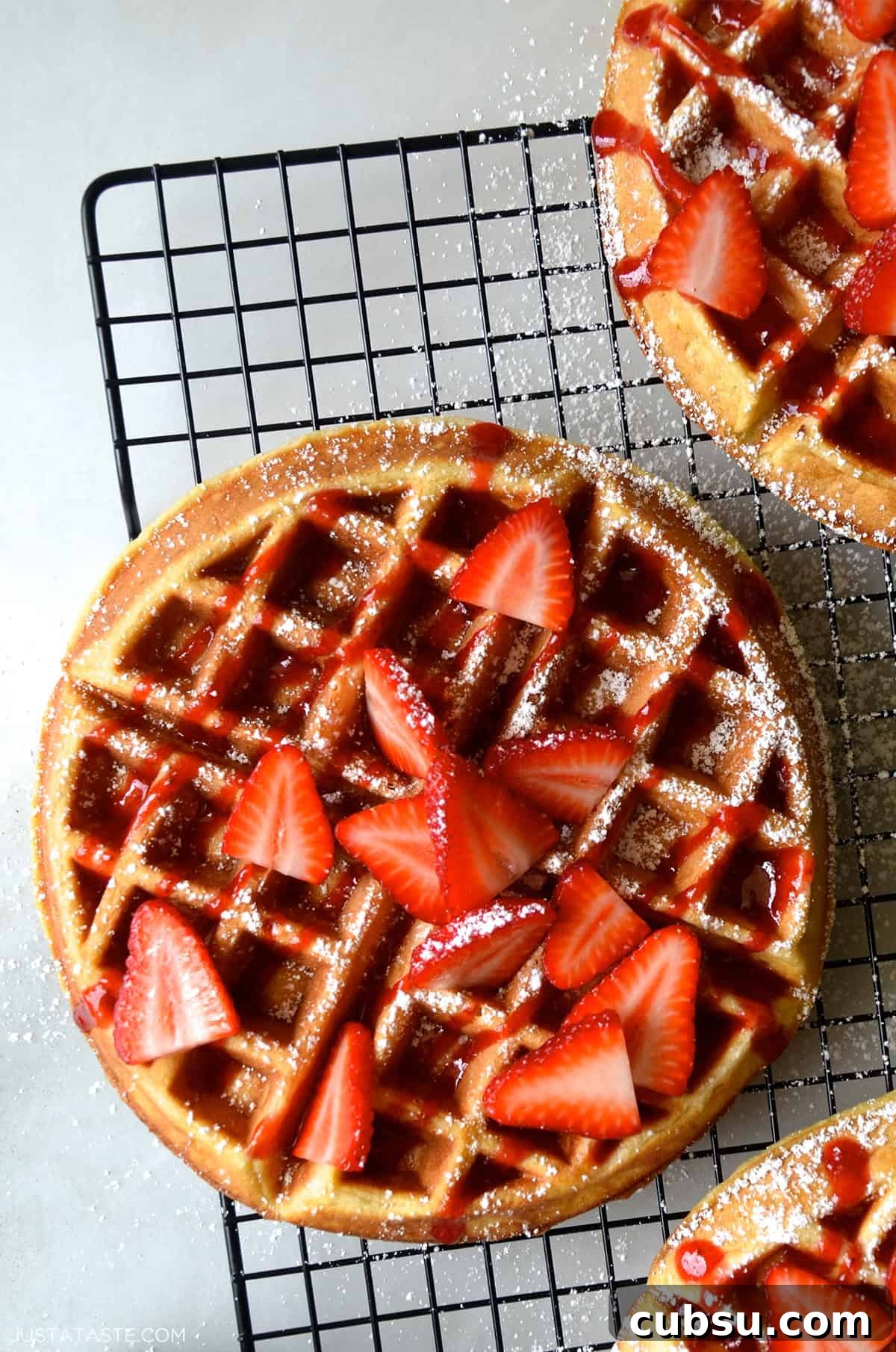 Crispy Belgian Waffles Recipe for Brunch and Weekend Mornings 2 A homemade waffle topped with maple syrup, fresh strawberries and a light dusting of powdered sugar on a wire rack.