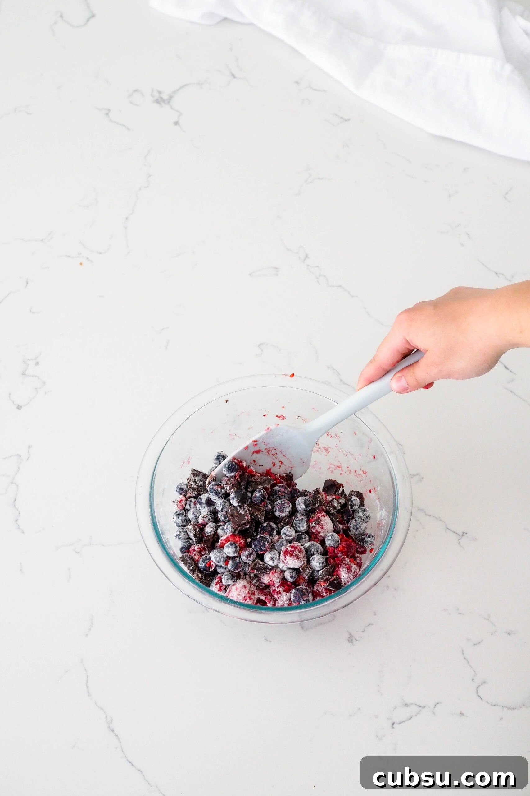 A hand mixes a glass bowl of berries, chocolate, and flour with a spatula.