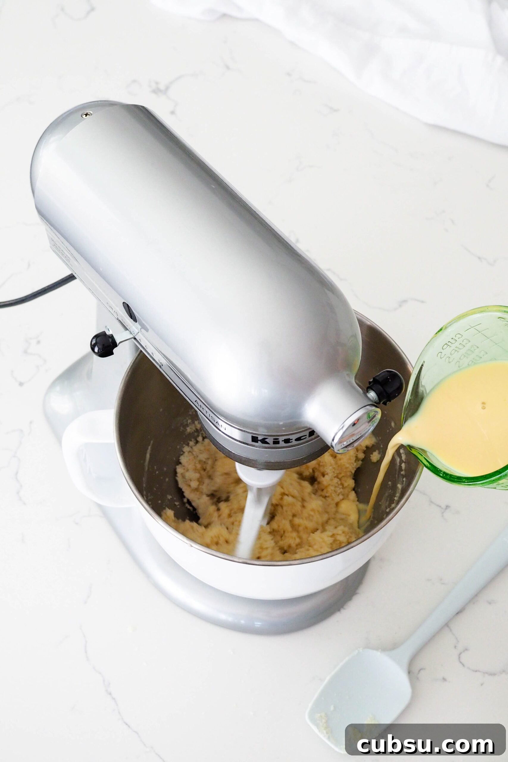 A spouted measuring cup pours wet ingredients into the bowl of a stand mixer.