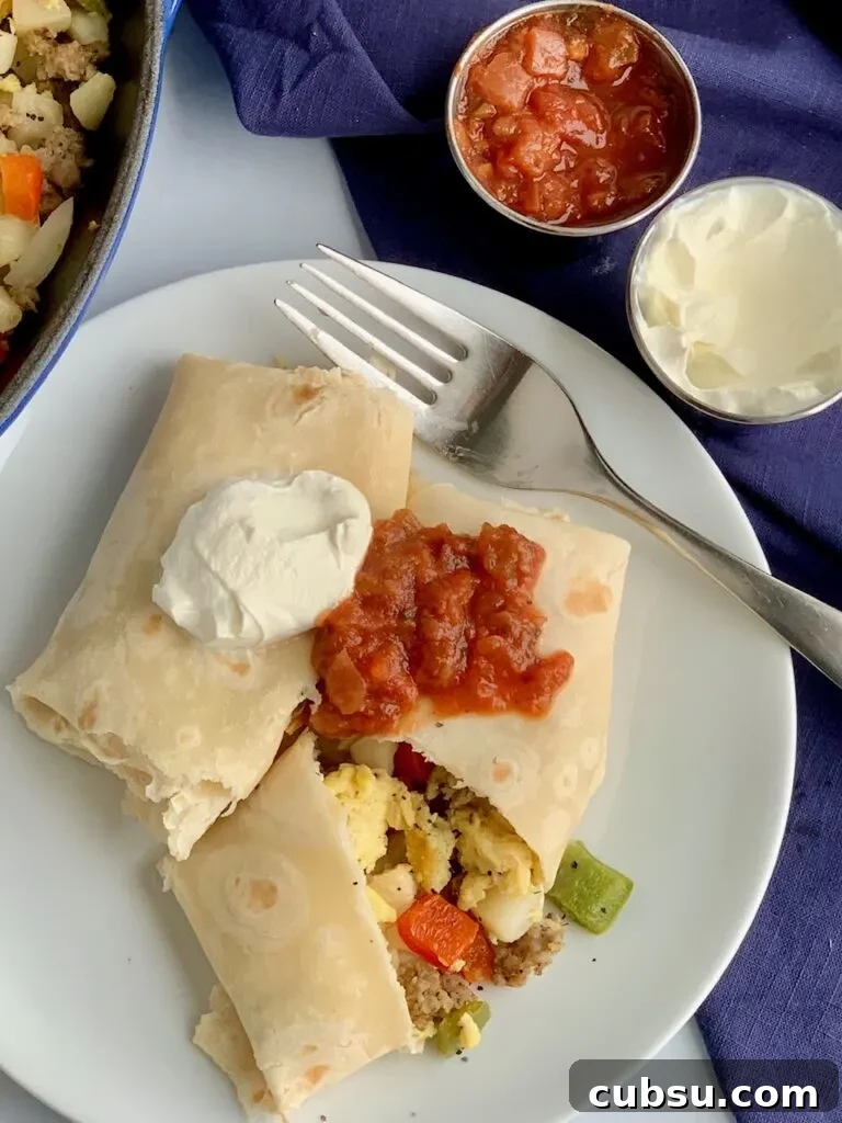 Make-Ahead Breakfast Burritos for Busy Mornings 7 A white plate with 2 large breakfast burritos on it. One is cut open and you can see eggs, bell peppers, sausage, and hash browns. They are topped with dairy free sour cream and a dollop of salsa