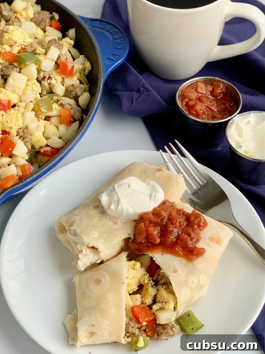 Make-Ahead Breakfast Burritos for Busy Mornings 8 A white plate with 2 large breakfast burritos on it. One is cut open and you can see eggs, bell peppers, sausage, and hash browns. They are topped with dairy free sour cream and a dollop of salsa.