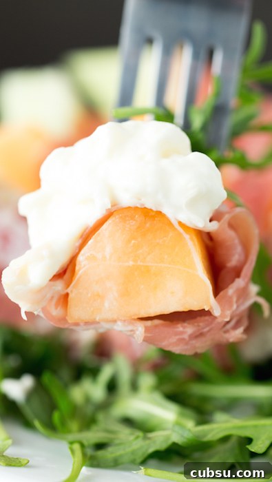 Prosciutto, Melon & Burrata Salad with Balsamic Glaze 4 A fork holding a bite sized piece of cantaloupe that's wrapped in prosciutto and has a bit of burrata on top.