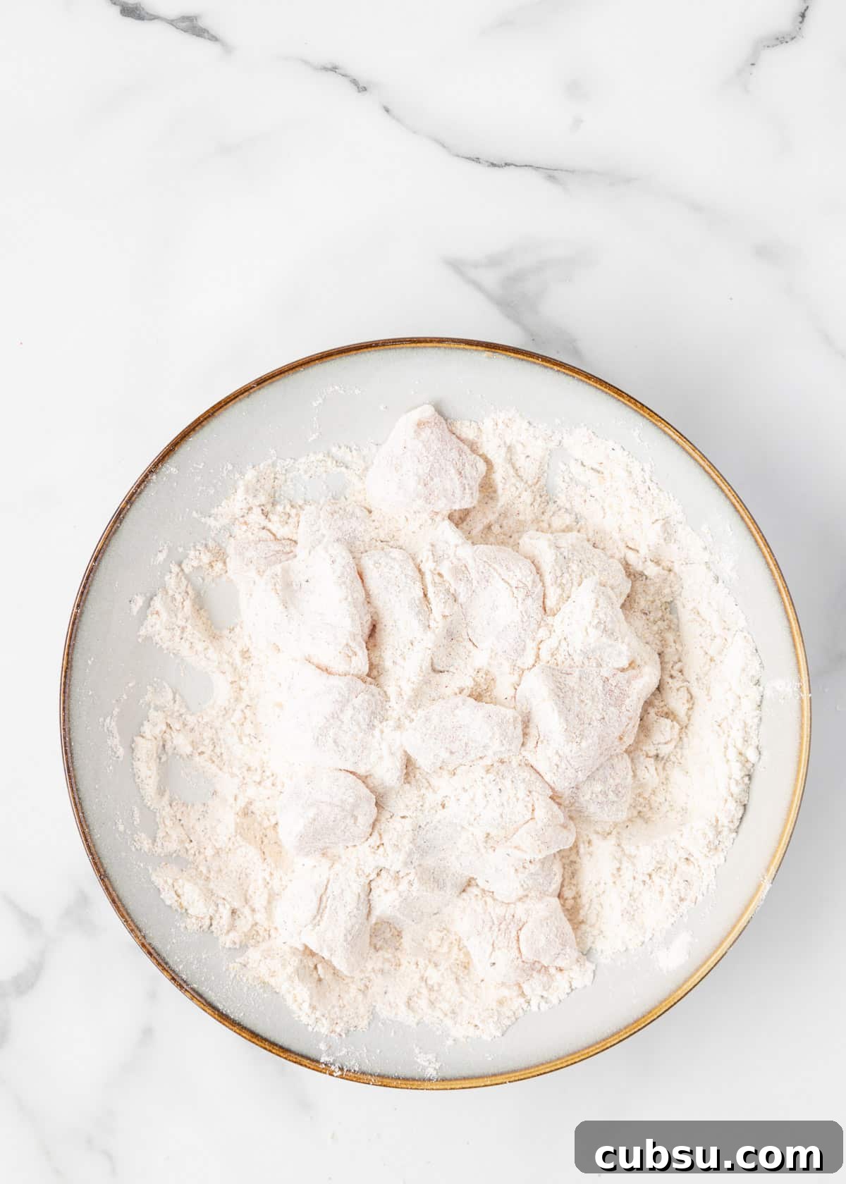 raw chicken cubes dredged in seasoned flour in a shallow bowl on a white marble board