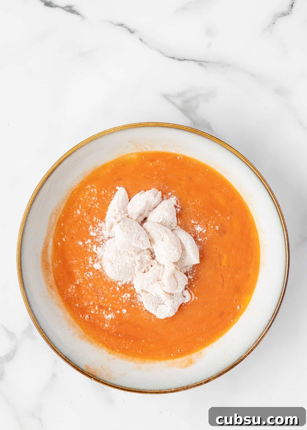 chicken tender pieces dredged in flour in a buffalo sauce egg wash in a shallow bowl on a white marble board