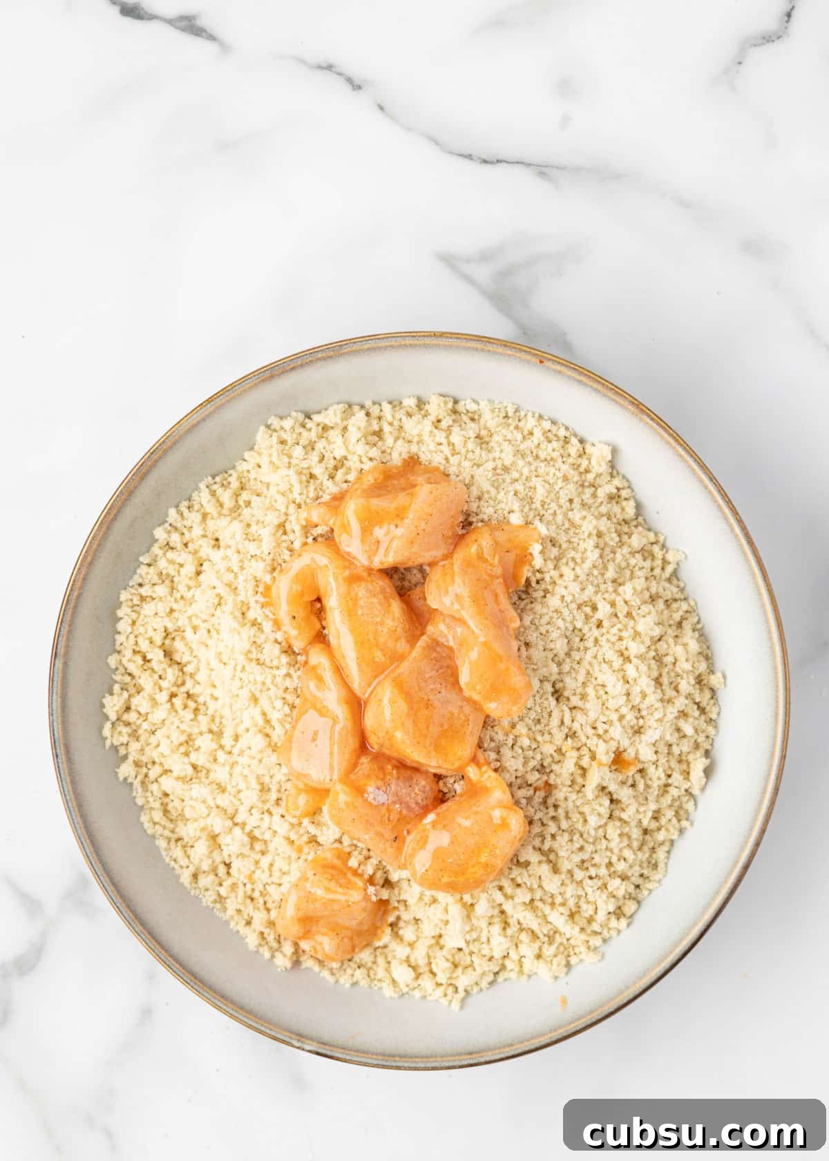 egg washed boneless chicken cubes in a bowl of seasoned panko breadcrumbs in a white marble board