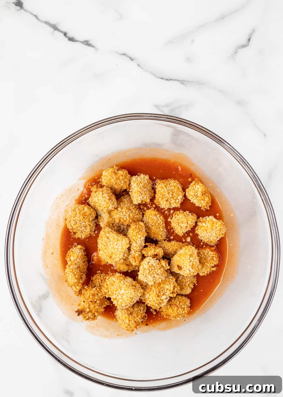 air fried buffalo chicken bites sitting on top of buffalo sauce, untossed, in a glass bowl on a white marble board