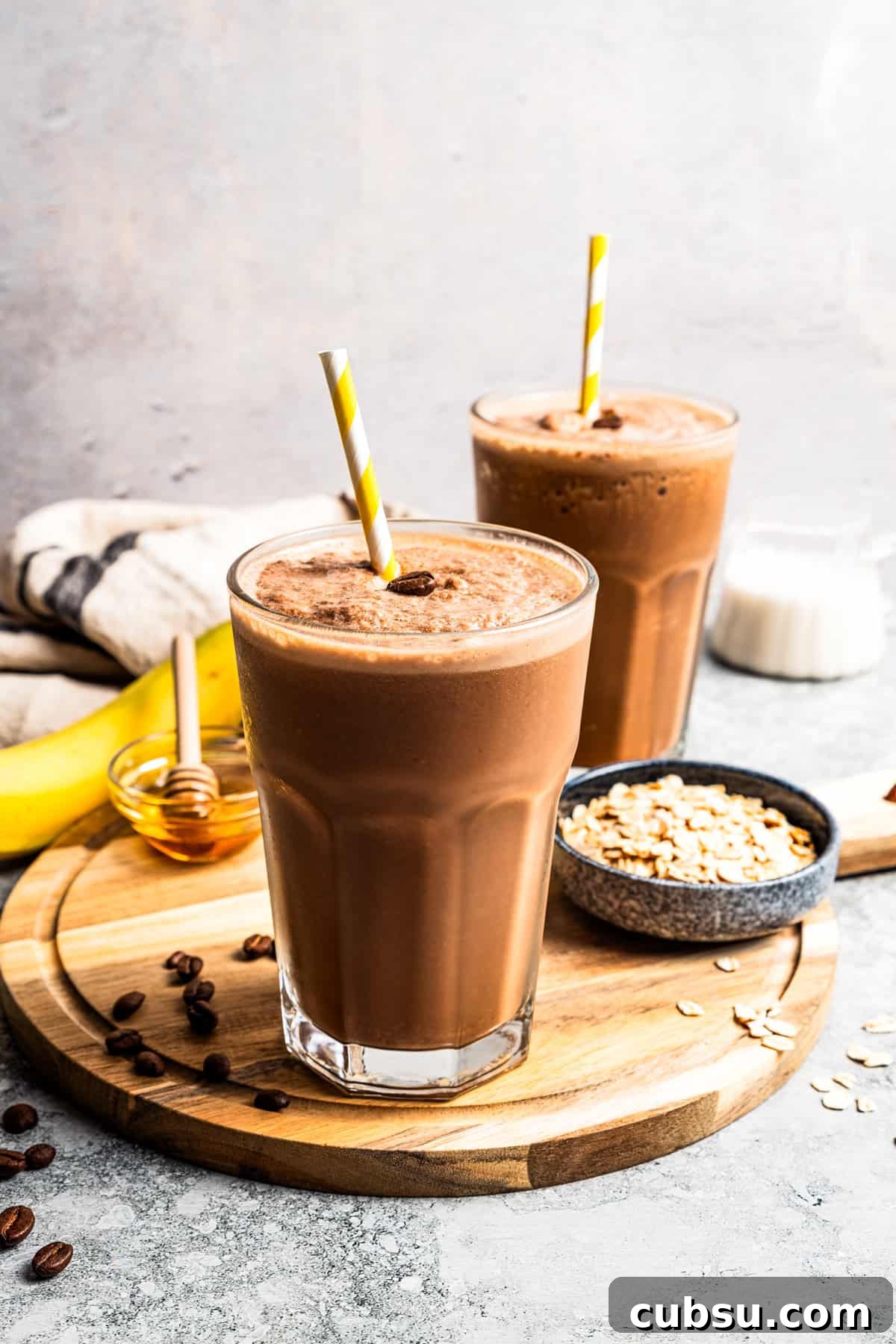This creamy coffee smoothie is a delicious and nourishing caffeine boost on busy mornings! Packed with wholesome oats, flaxseed, and bananas. A coffee smoothie in a tall glass with a straw and a second smoothie next to a bowl of oats in the background.