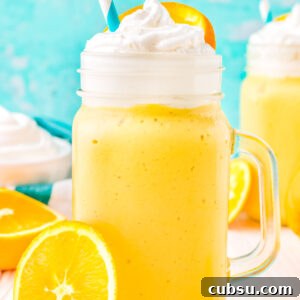 An orange milkshake in a clear glass garnished with whipped cream, a paper straw, and an orange slice.
