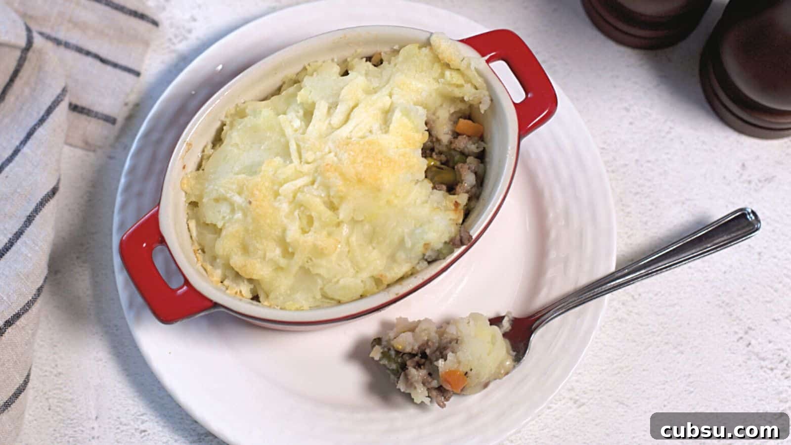 17 Timeless Grandma Recipes You Need to Try Now 12 Shepherd's pie in a red ceramic dish with a spoon, showcasing layers of mashed potatoes and minced meat with vegetables on a white tablecloth.