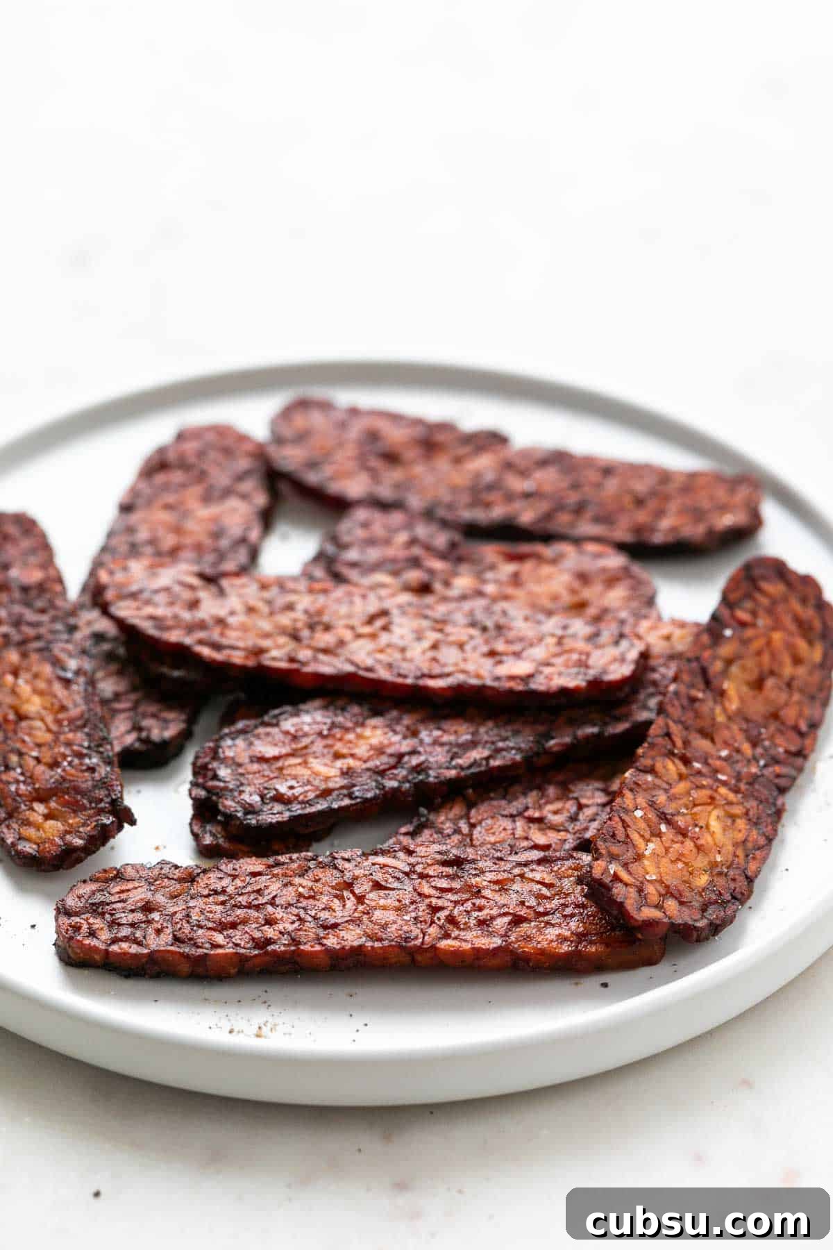 Crispy Tempeh Bacon Recipe: Smoky Vegan Bacon Alternative 2 White plate with slices of tempeh bacon.