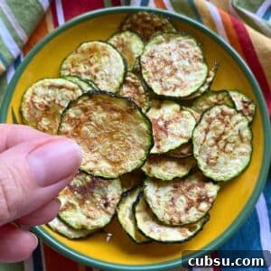 Finger holding a zucchini chip cooked in the microwave over a yellow plate with more zucchini chips.