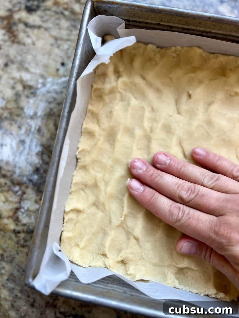 Sourdough Sugar Cookie Bars Recipe — Buttery Soft Cookie Bars 6 A hand pressing sourdough sugar cookie bars into a parchment-lined baking pan.