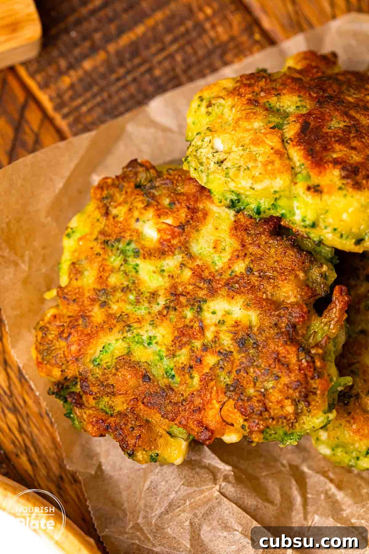 Close up of crispy pan fried broccoli fritters stacked on parchment paper, showing a golden brown crust and green broccoli pieces inside.