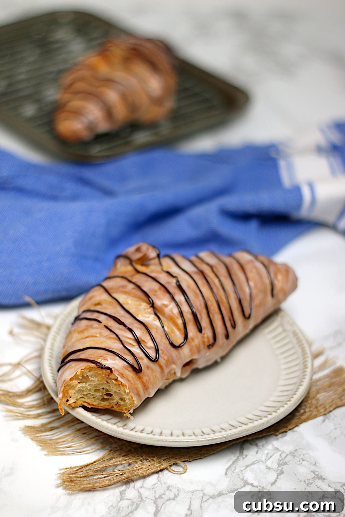 A chocolate drizzled glazed croissant with a bite missing on a small white plate.