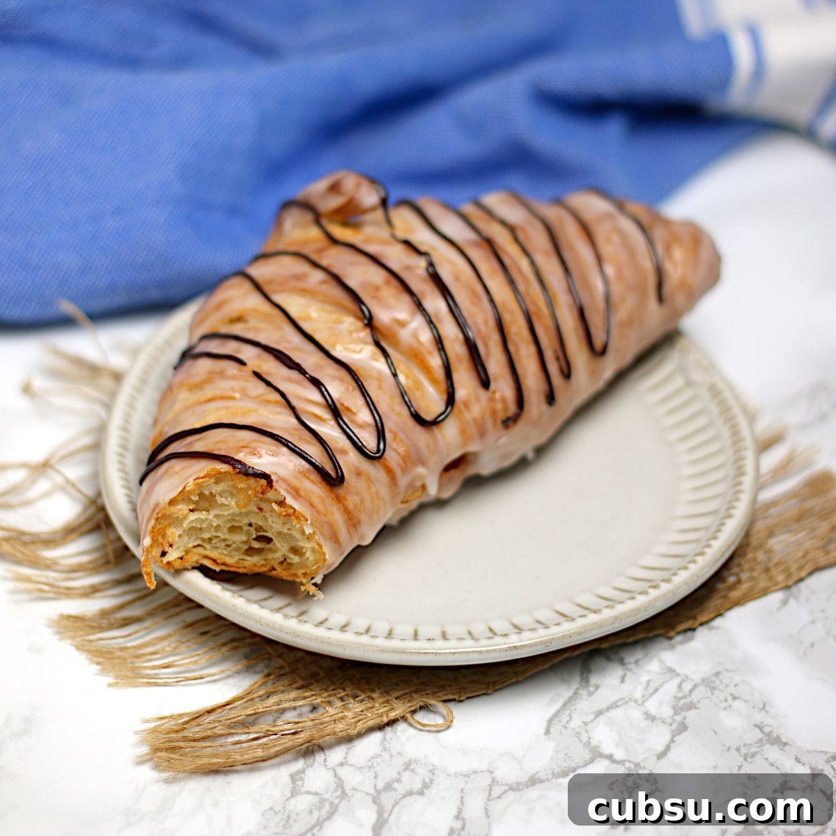 A chocolate drizzled glazed croissant with a bite missing on a small white plate.