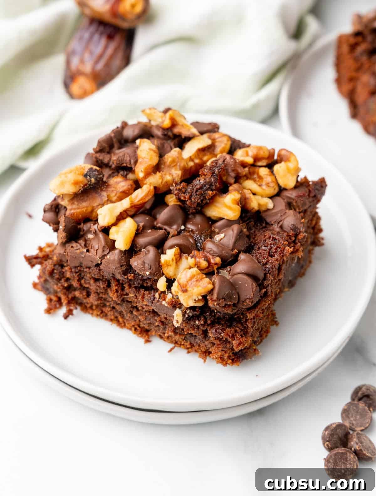 Decadent Chocolate and Date Cake Recipe for Moist Loaves 2 Square of chocolate chip walnut date cake on stack of two white plates. White surface, kitchen towel, loose dates around.