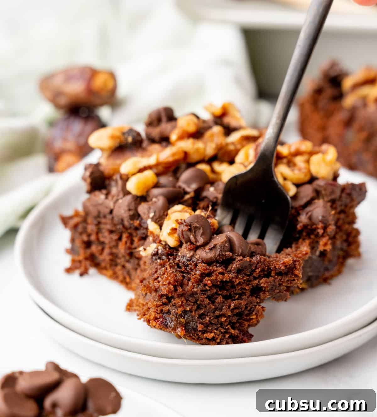 Decadent Chocolate and Date Cake Recipe for Moist Loaves 12 Dark fork with bite of chocolate walnut date cake on a white plate with rest of serving.