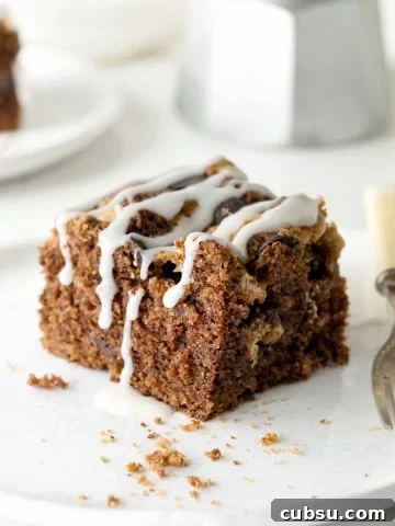 Single glazed chocolate cake square on a white plate, a fork. White background with metal coffee pot and more cake.