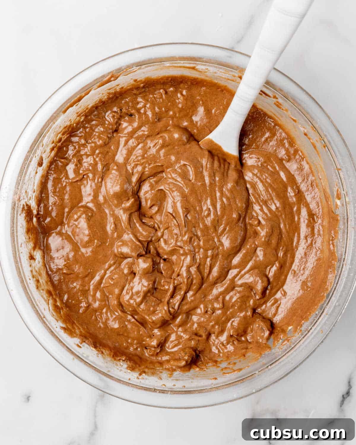 Decadent Chocolate and Date Cake Recipe for Moist Loaves 9 Chocolate date cake batter in a glass bowl with a spatula on a marble surface.